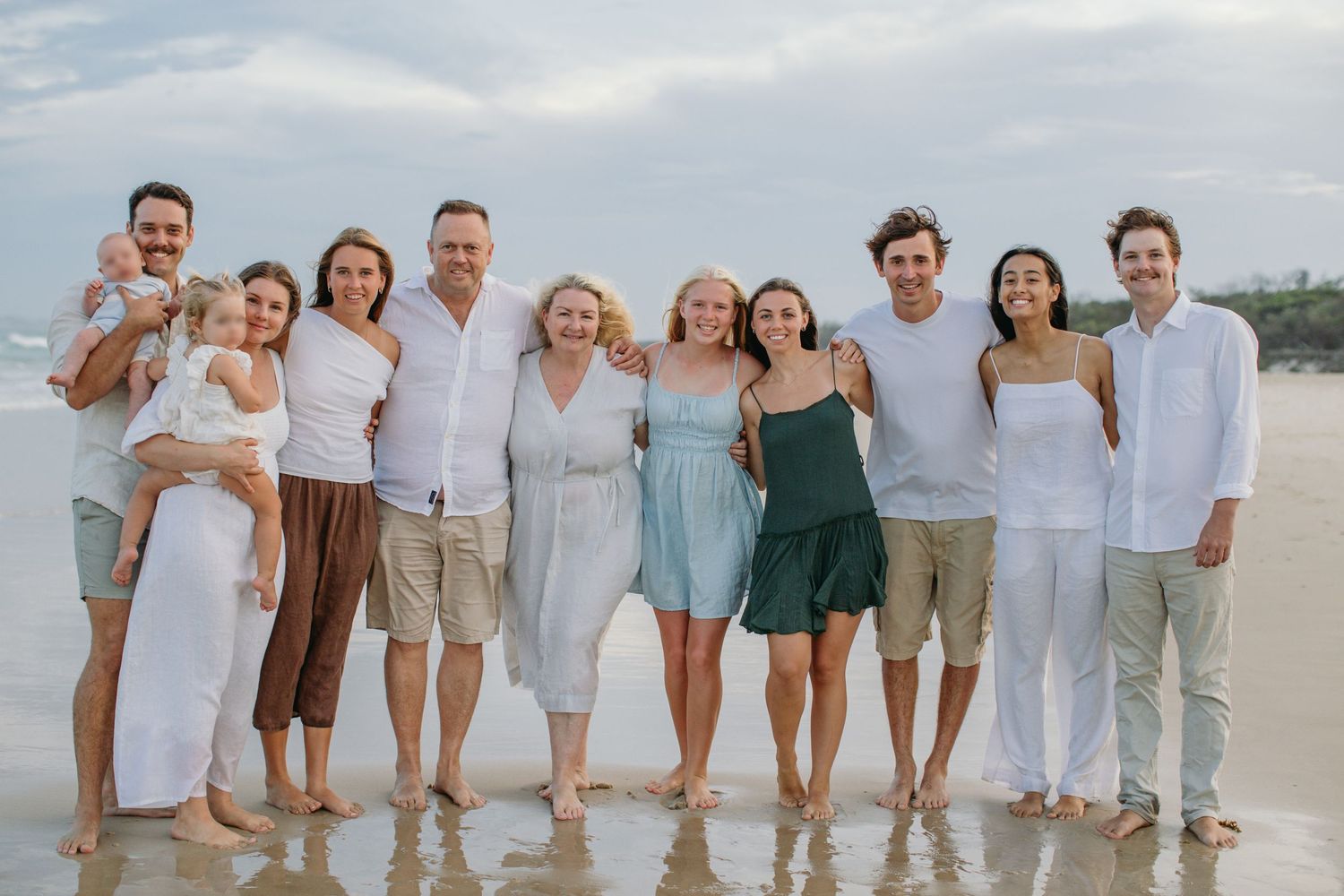 Large family group smiling together on a sandy beach, wearing coordinated white and neutral outfits on a cloudy day.