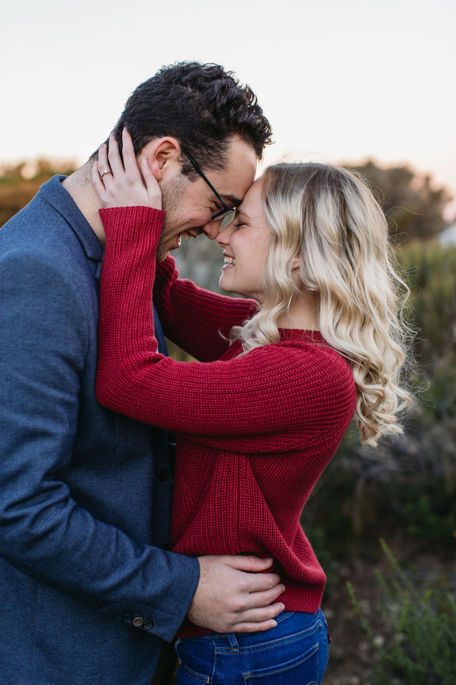 A couple in red and blue sweaters share a romantic forehead-to-forehead moment outdoors at sunset.