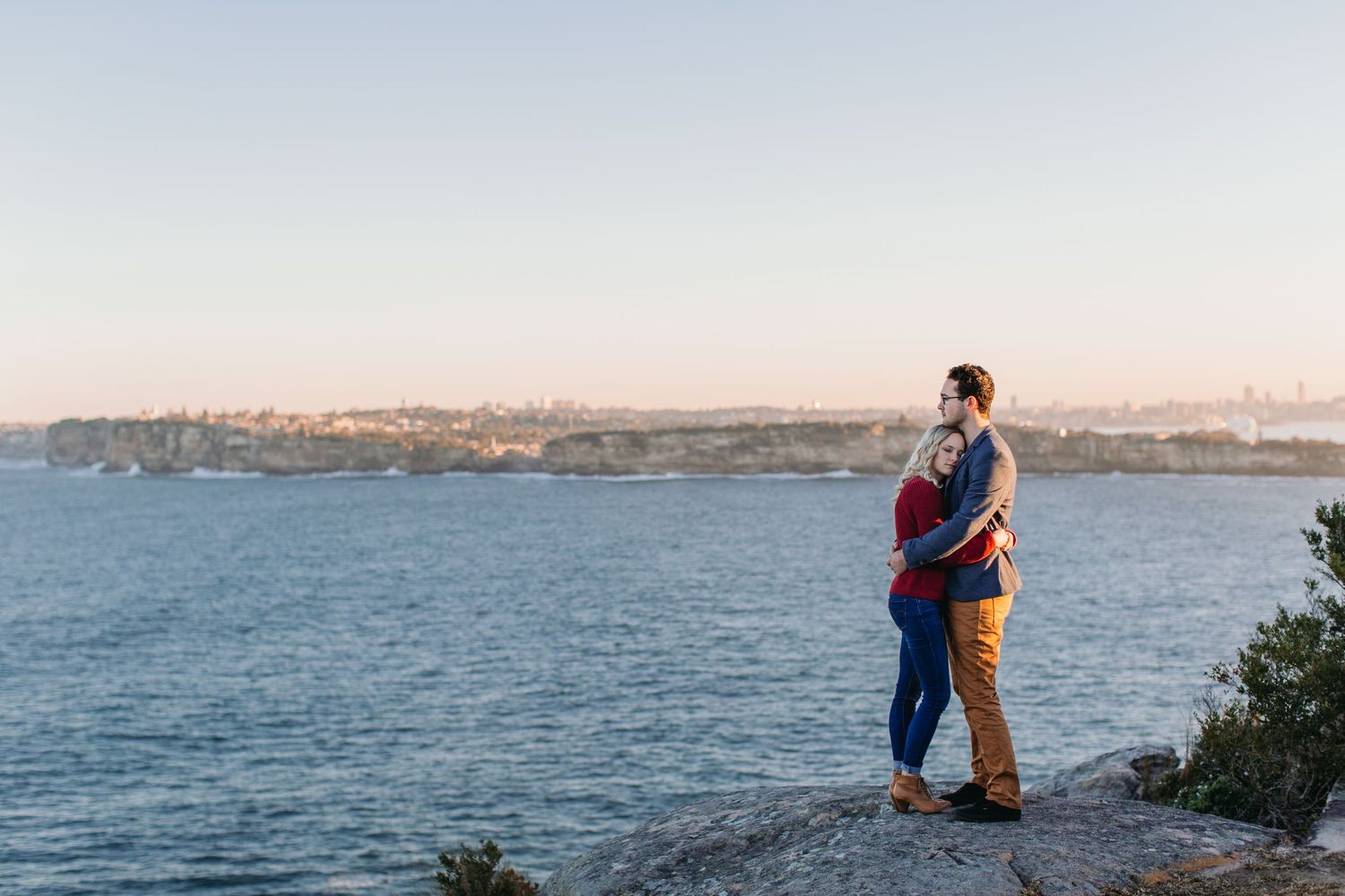 Couple embracing on rocky cliff overlooking serene blue harbor with city skyline at golden hour sunset.