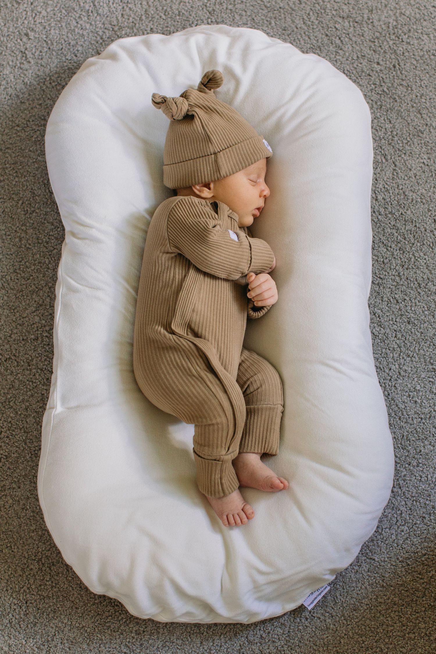 Newborn baby sleeping peacefully in a white snuggle nest, wearing a tan ribbed romper and matching knotted beanie hat.