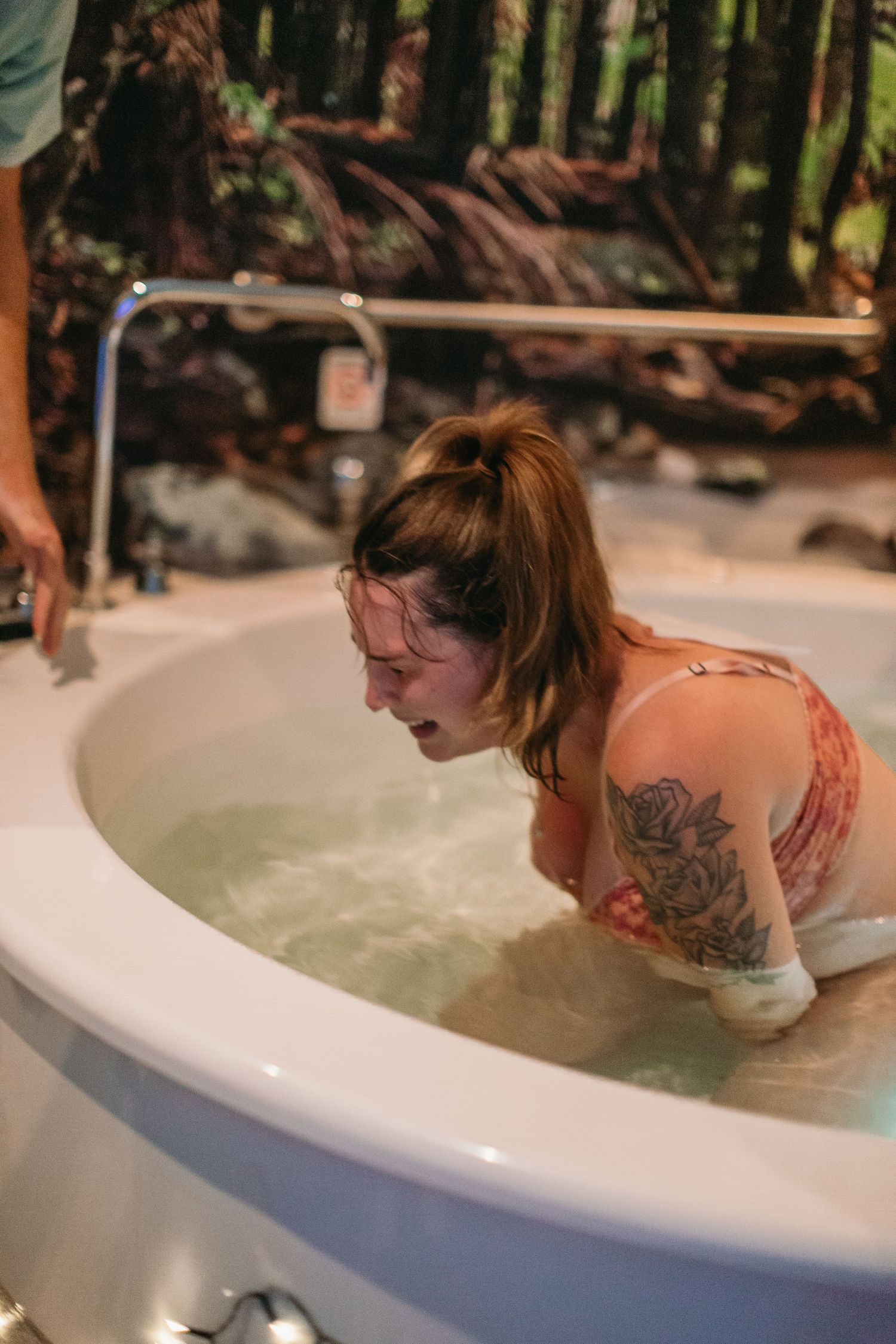 Tattooed woman in labor leaning over a birthing tub during water birth.