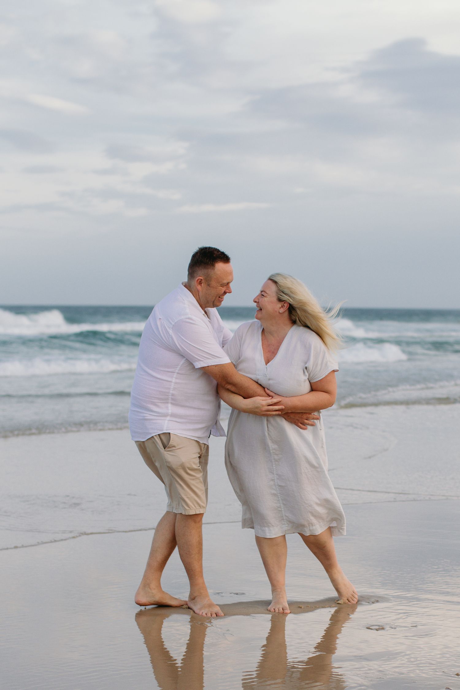 Couple in white outfits dancing barefoot on a sandy beach with ocean waves in the background.