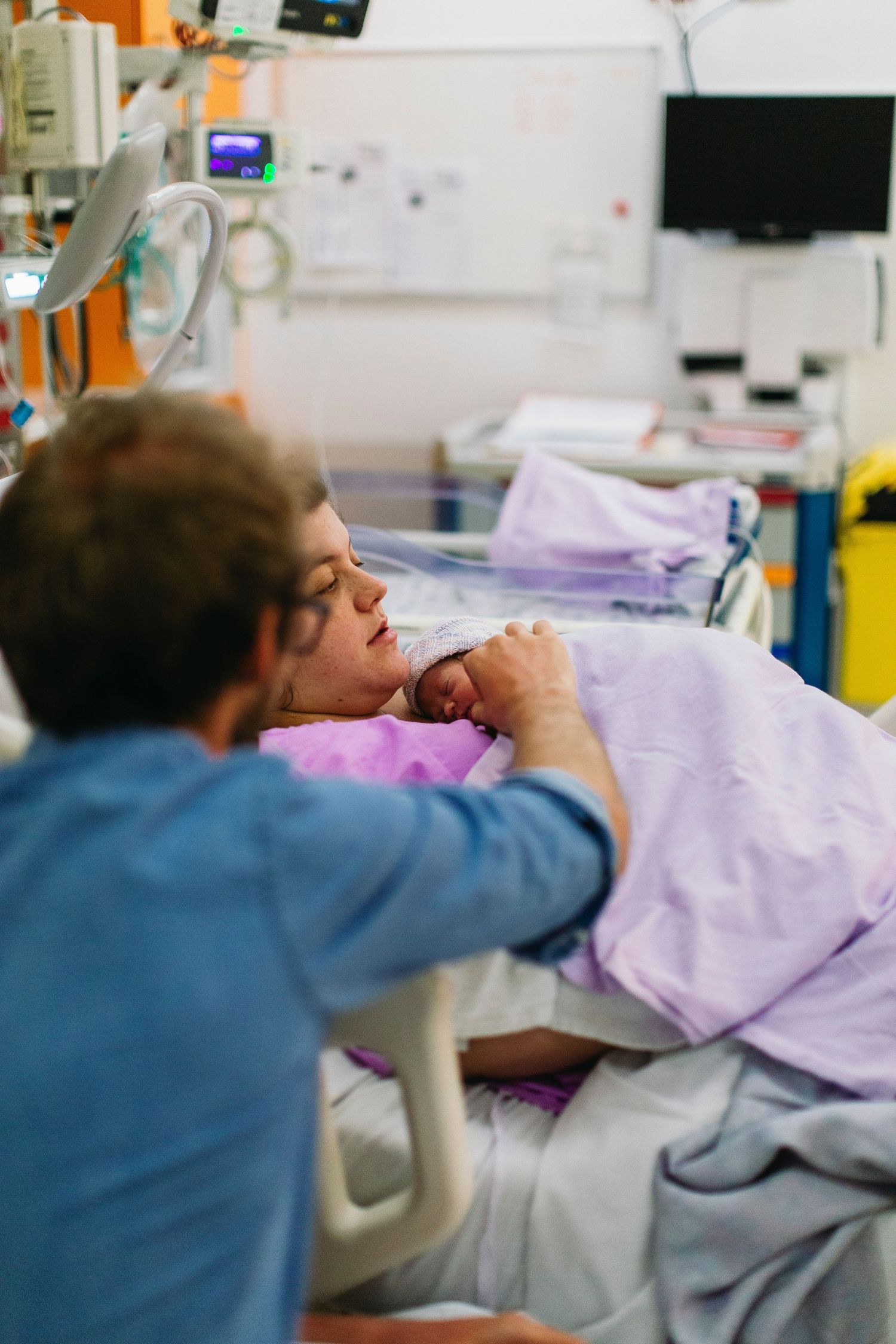 New mother in hospital bed holds newborn baby while partner leans in close during emotional post-birth moment.