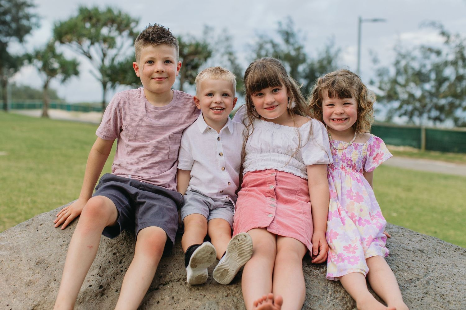 Four children sitting together on a rock outdoors, smiling happily in a park setting on a cloudy day.