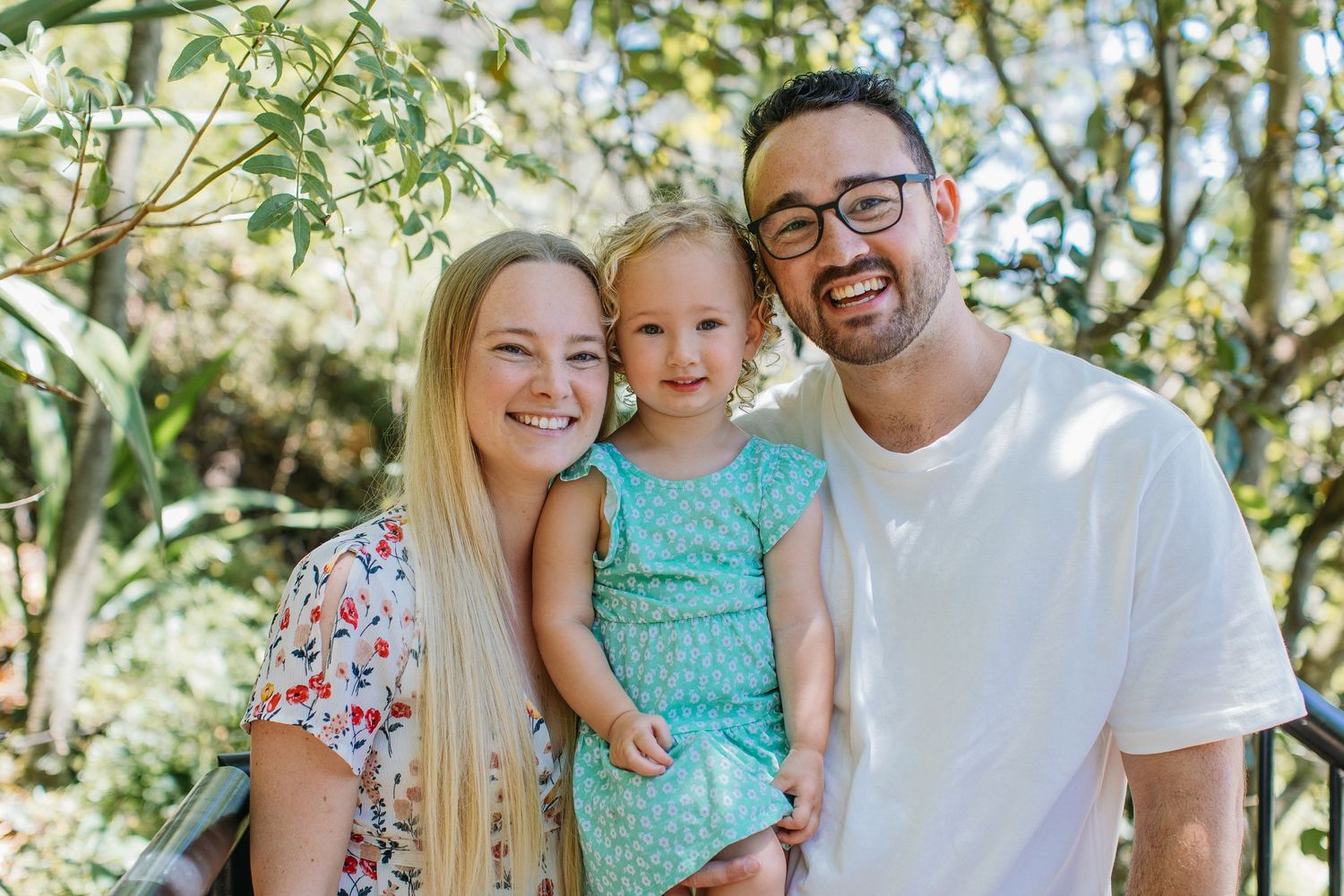 Happy family portrait with smiling parents and toddler girl in teal dress outdoors among green trees.