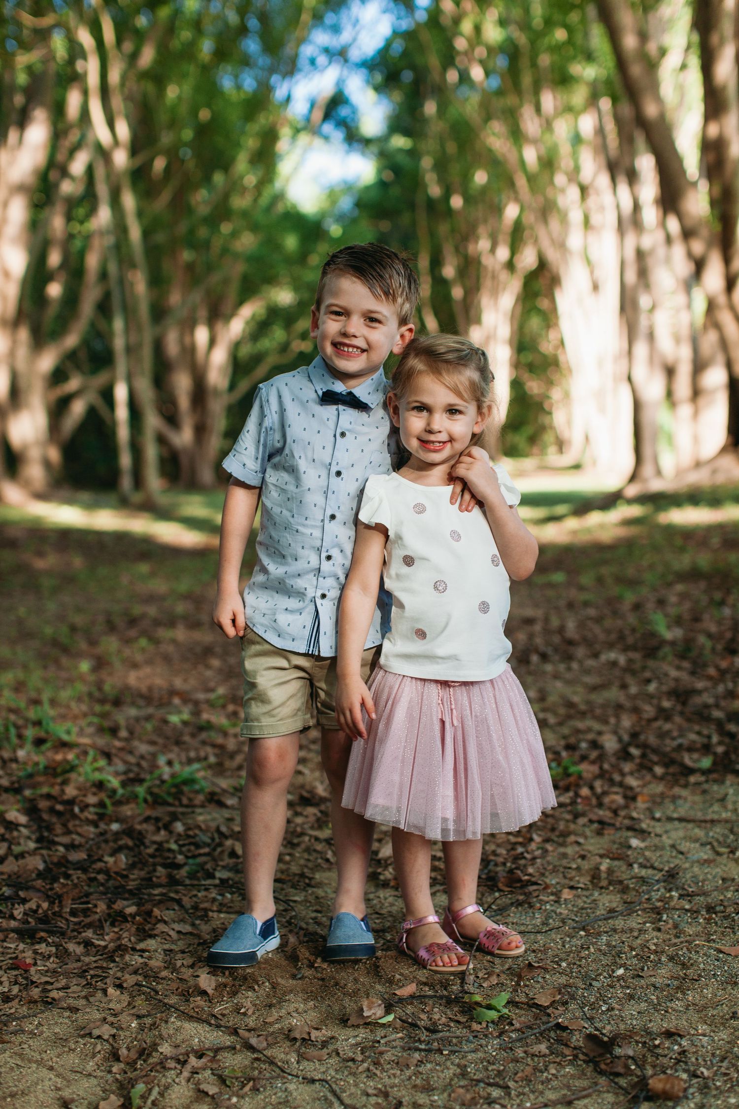 Two young children, a boy in a blue shirt and a girl in a pink tutu skirt, pose together on a tree-lined path.