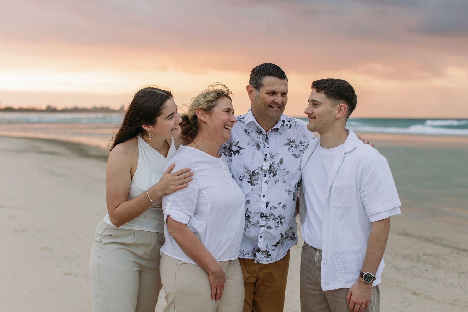 Family of four laughing together on a sandy beach at sunset with colorful pink and purple skies.