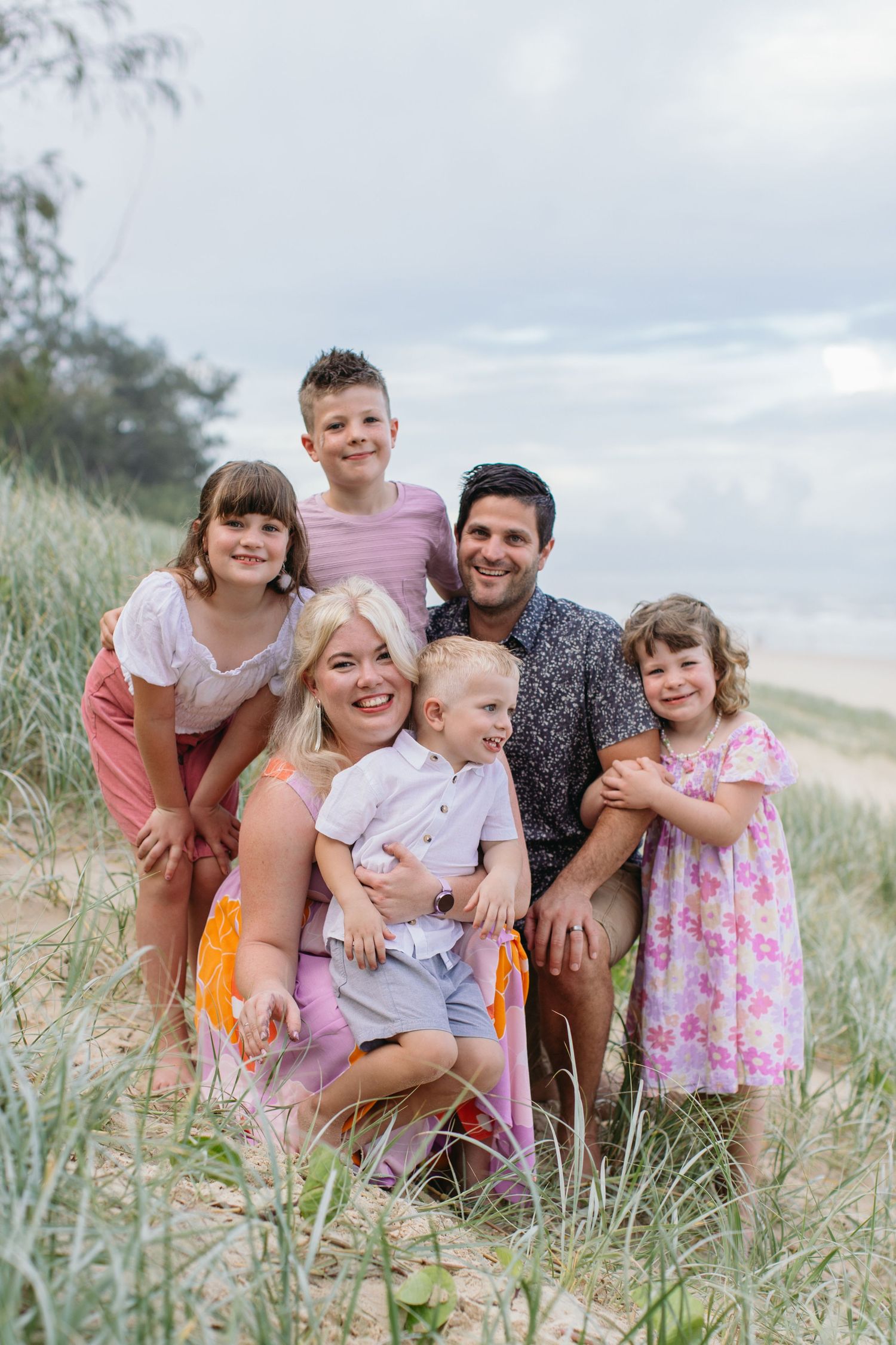 Happy family of six posing together on sandy beach dunes with ocean backdrop on cloudy day.