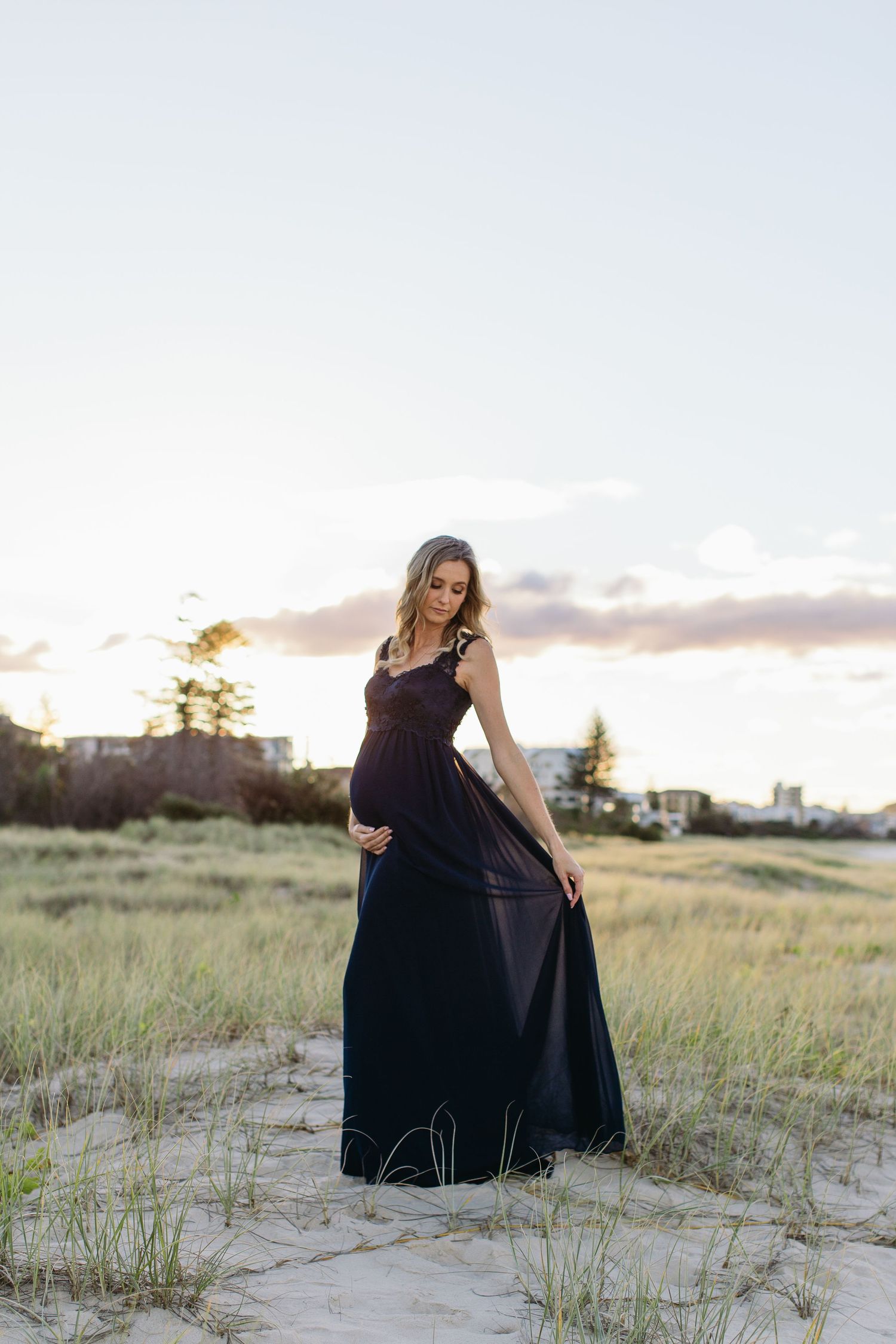 Pregnant woman in dark maxi dress standing on sandy beach dunes at sunset, cradling her baby bump.