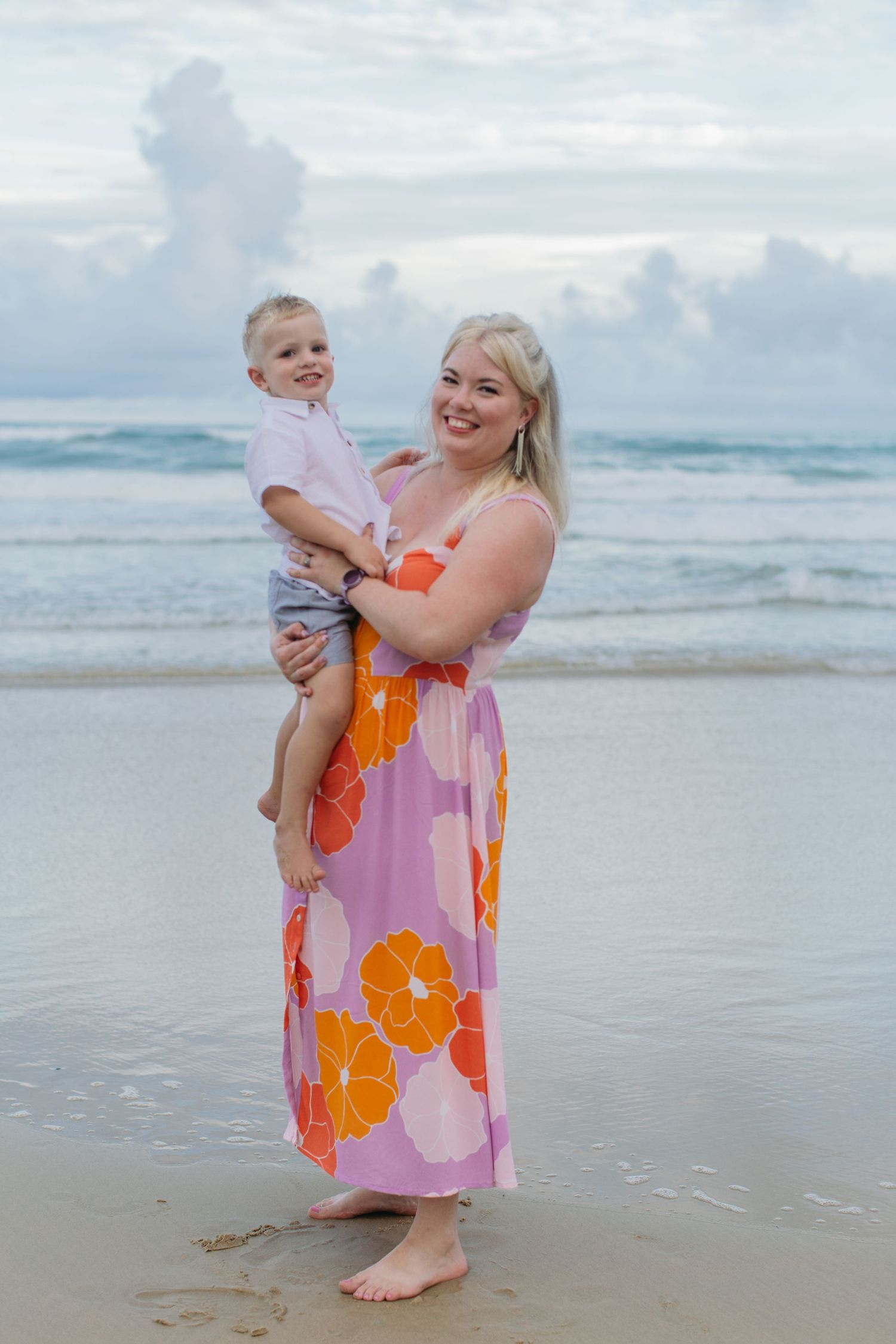 A blonde mother in a floral maxi dress holds her young son on a beach with ocean waves behind them.