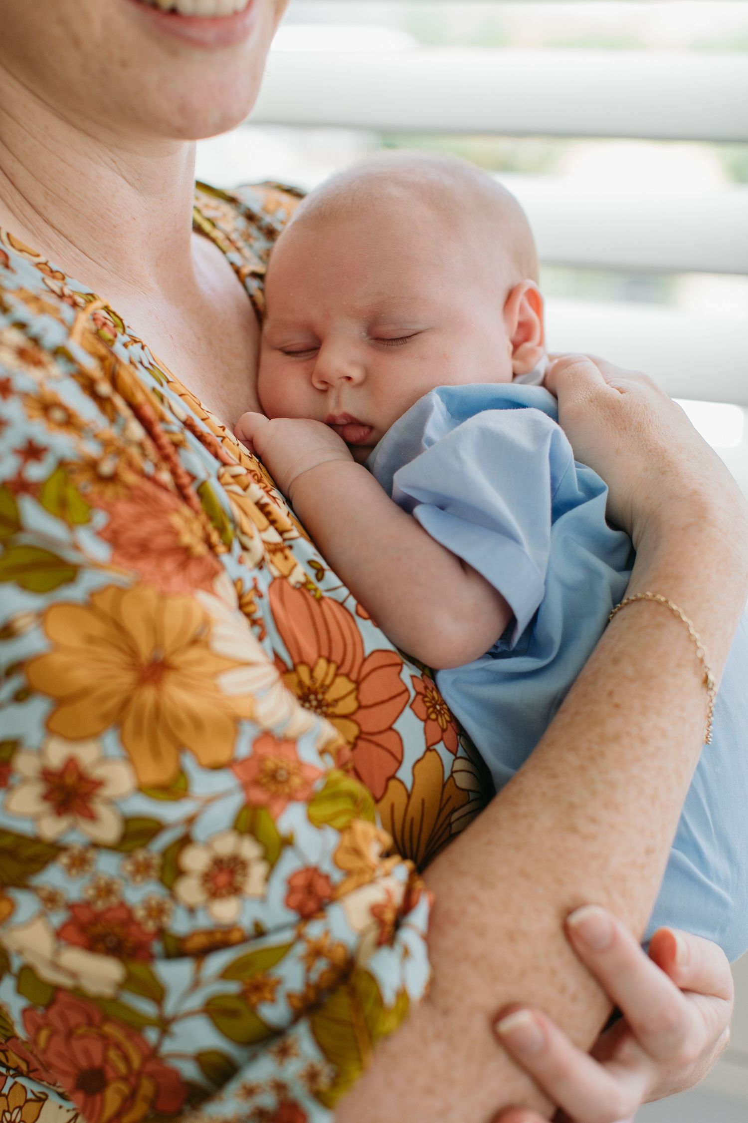 A sleeping newborn baby in blue outfit cradled in the arms of a smiling woman wearing a floral dress.