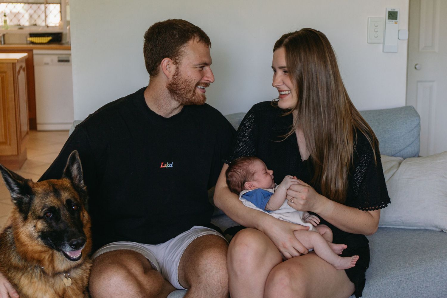 A couple sits on a couch smiling at their newborn baby while a German Shepherd dog sits beside them.