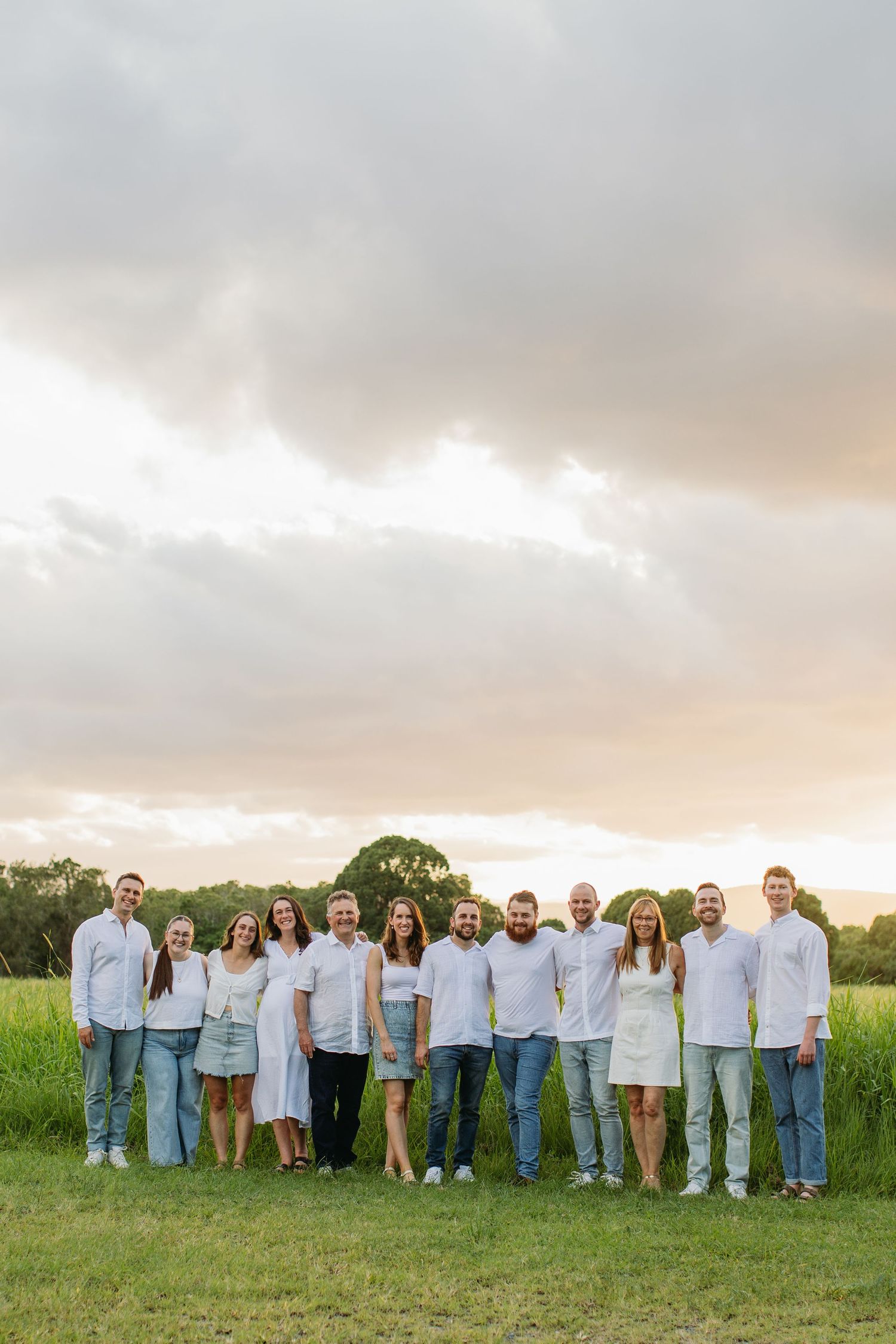 Large family group portrait outdoors in a green field at sunset, wearing coordinated white tops and jeans.