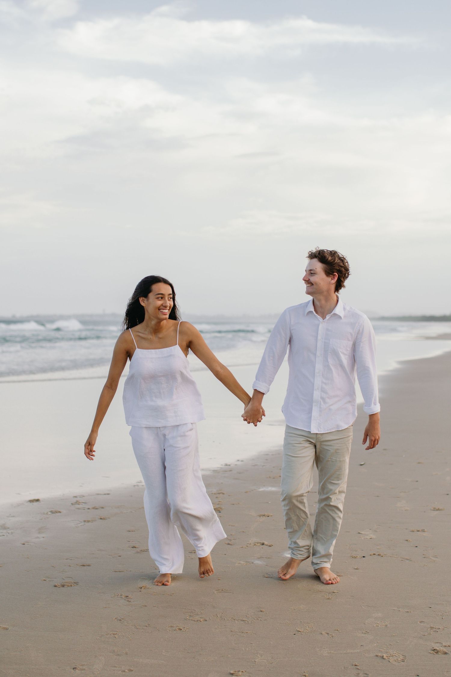 A happy couple walks hand-in-hand along a sandy beach, both wearing white outfits, with ocean waves in the background.