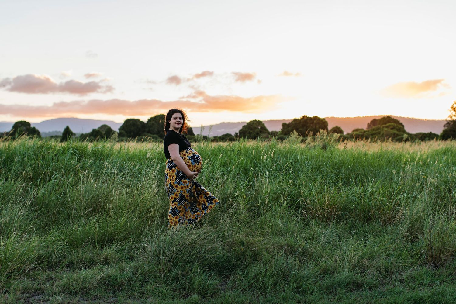 Pregnant woman in floral skirt standing in lush green field at golden sunset with mountains in background.