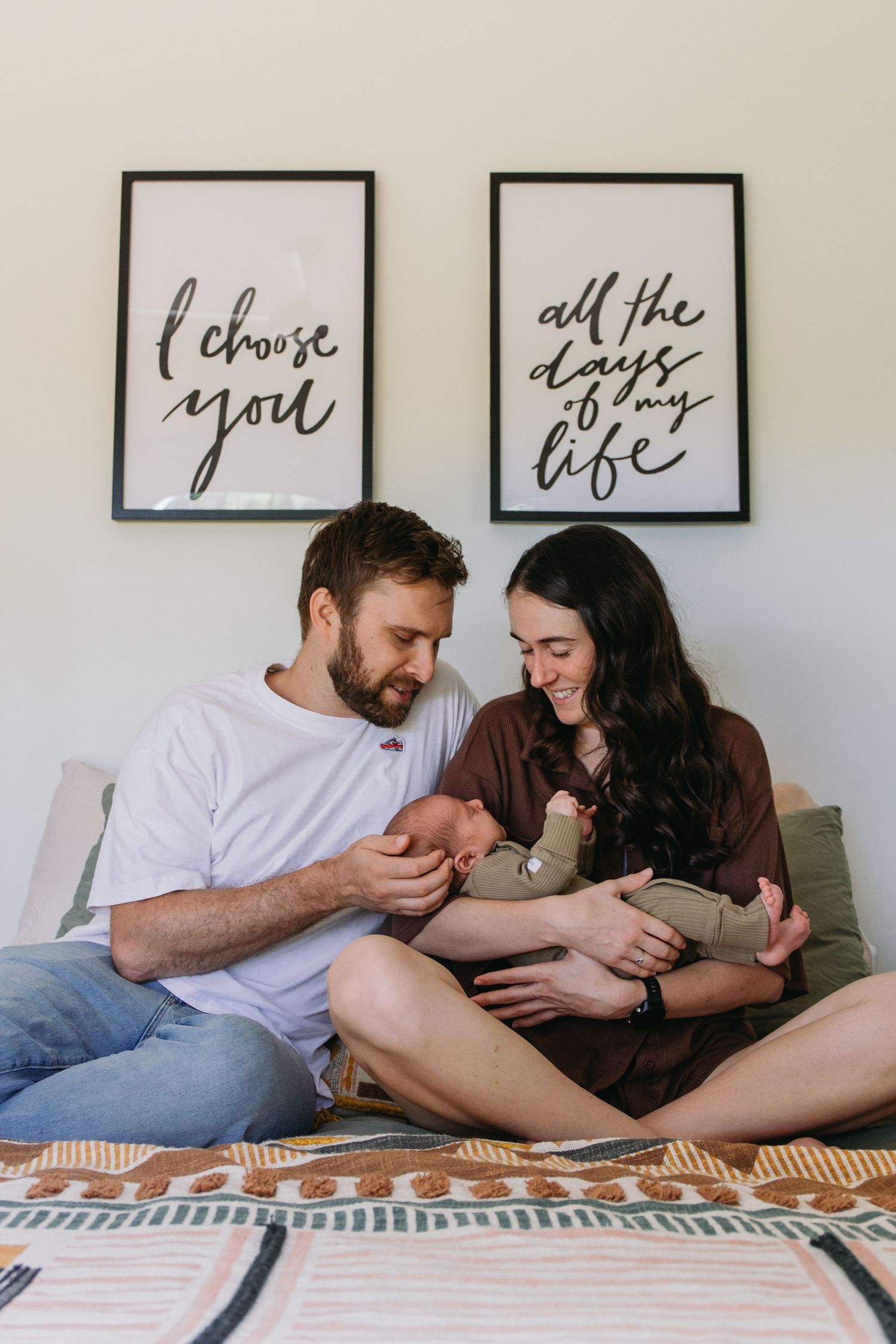 Happy couple sitting cross-legged on bed holding newborn baby, with romantic wall art reading I choose you all the days of my life.