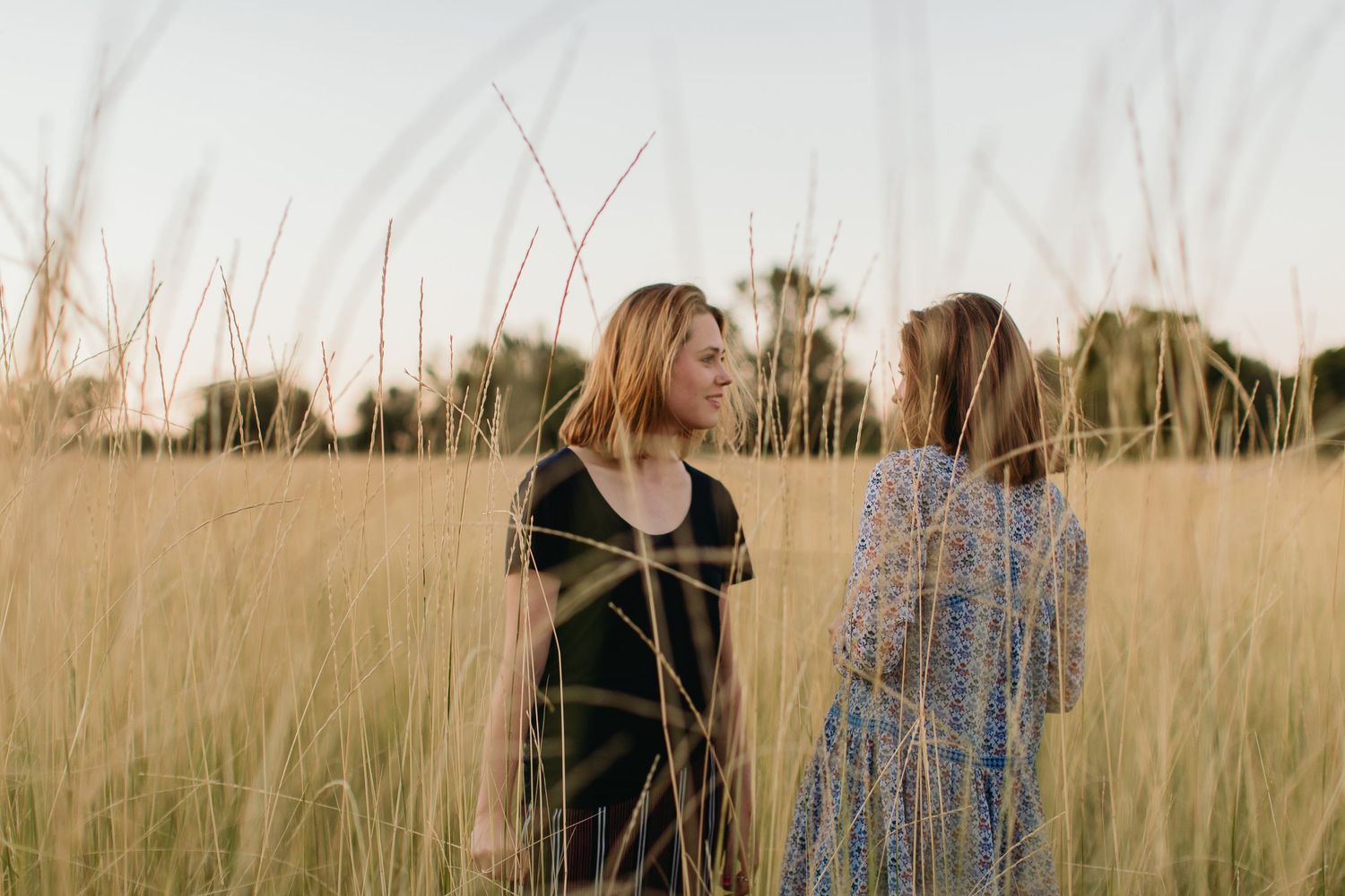 Two women standing in tall golden grass field having a conversation at sunset.