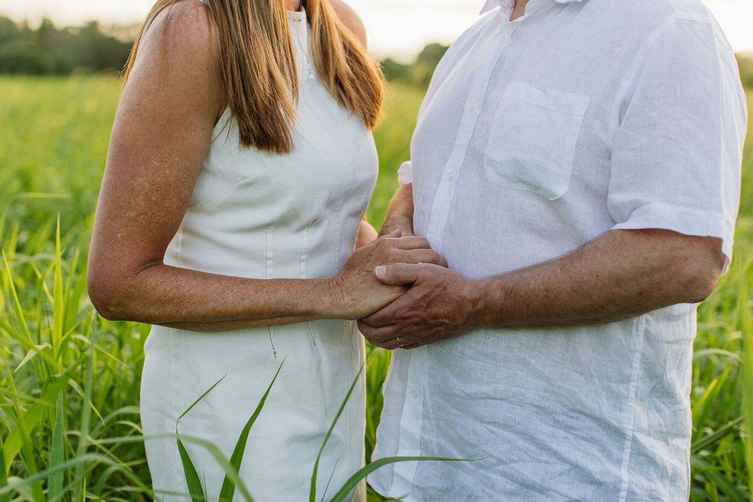 Couple holding hands in white outfits standing in a lush green field during golden hour.