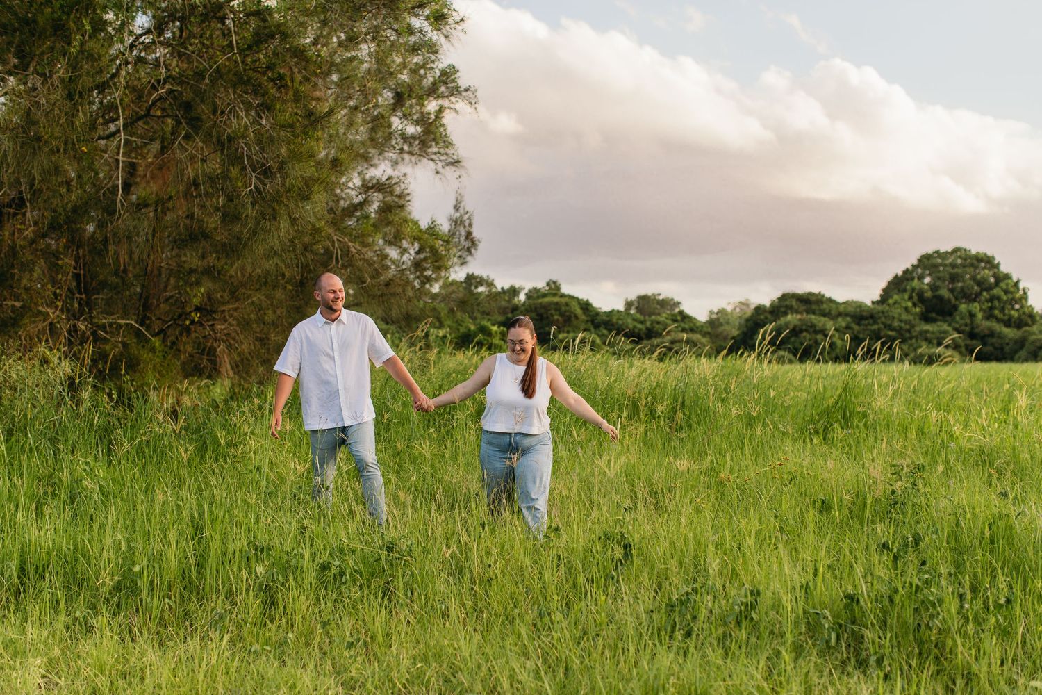Couple holding hands walking through lush green meadow with trees in background on cloudy day.