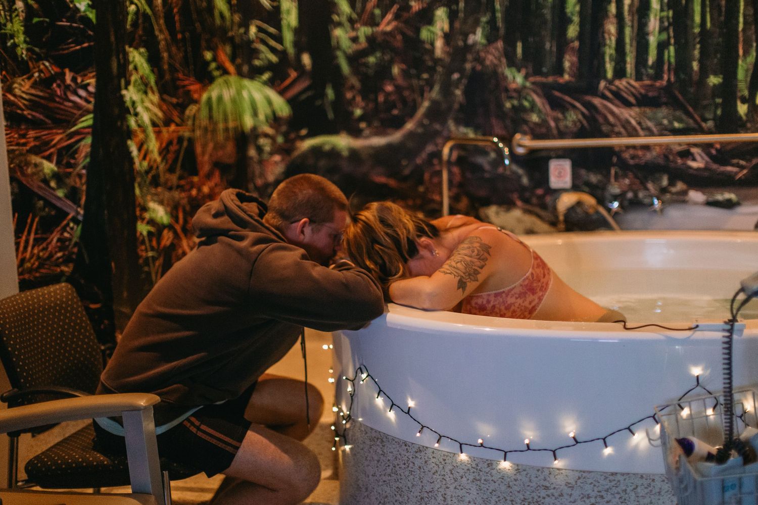 A couple shares an intimate moment at a fairy light-adorned birthing tub with a forest mural backdrop.