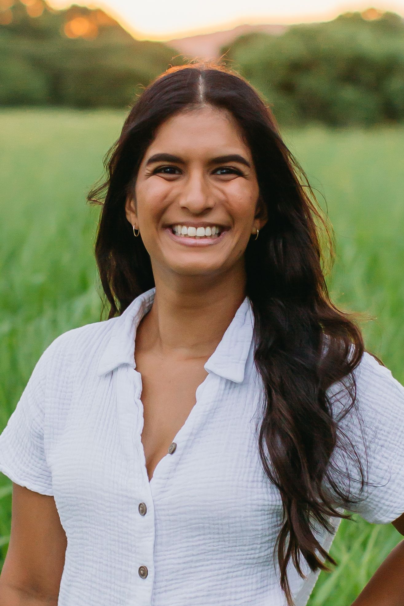 Smiling young woman with long dark hair wearing a white button-up shirt standing in a lush green field at sunset.