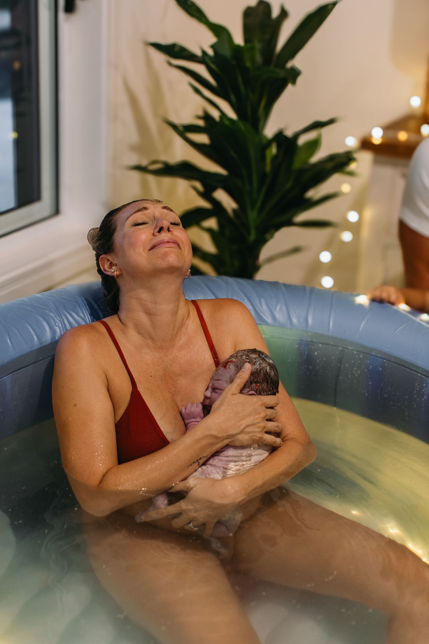 Tearful new mother embracing her newborn baby while sitting in a home water birth pool.