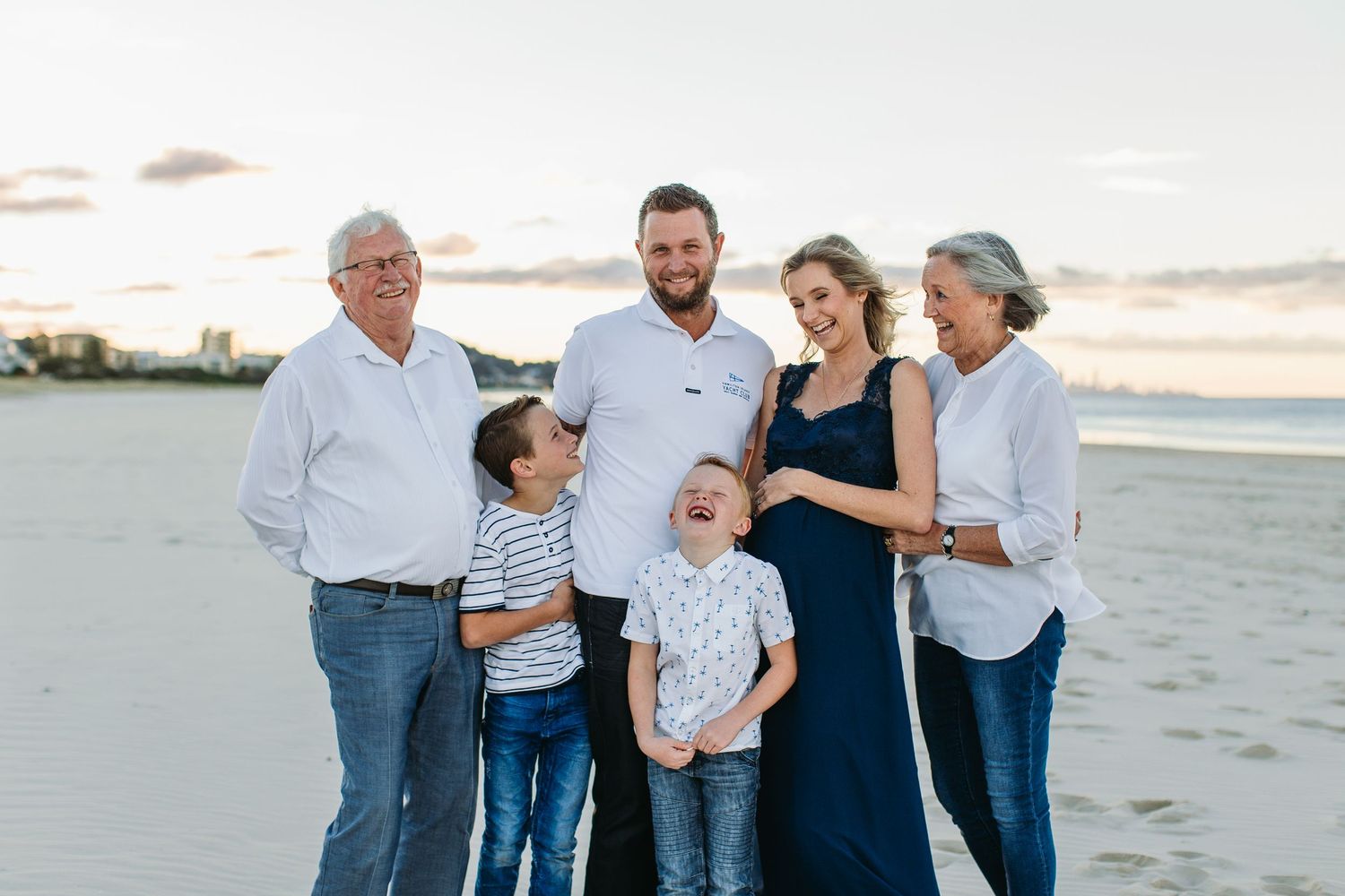 Multi-generational family of six smiling together on a sandy beach at sunset, including grandparents, parents, and two young boys.