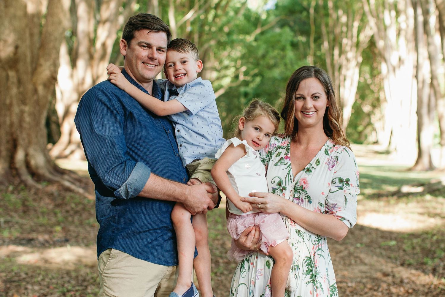 Happy family of four posing outdoors among tall trees, parents holding two young children, smiling in natural light.