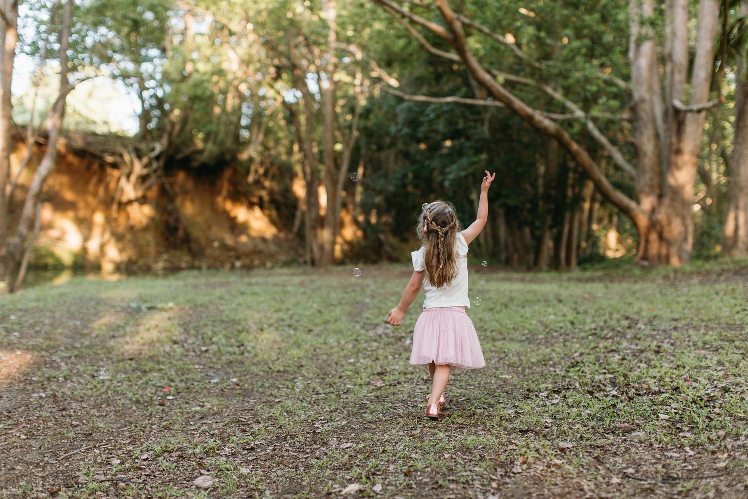 A young girl in a pink tutu skirt walks toward a lush green forest, reaching up with one hand.