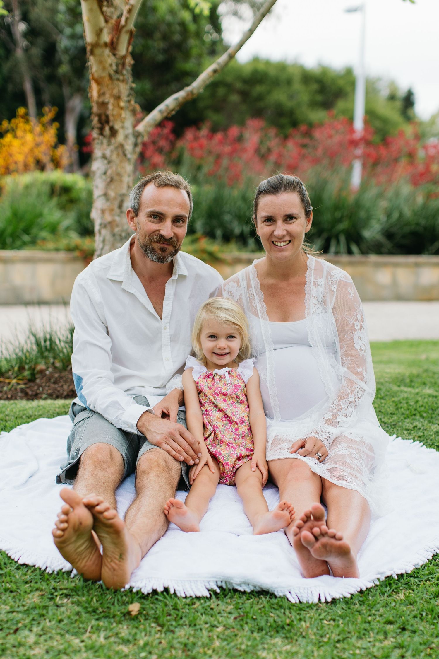 A happy family of three sits barefoot on a white blanket outdoors, with a pregnant mother, father, and young daughter.