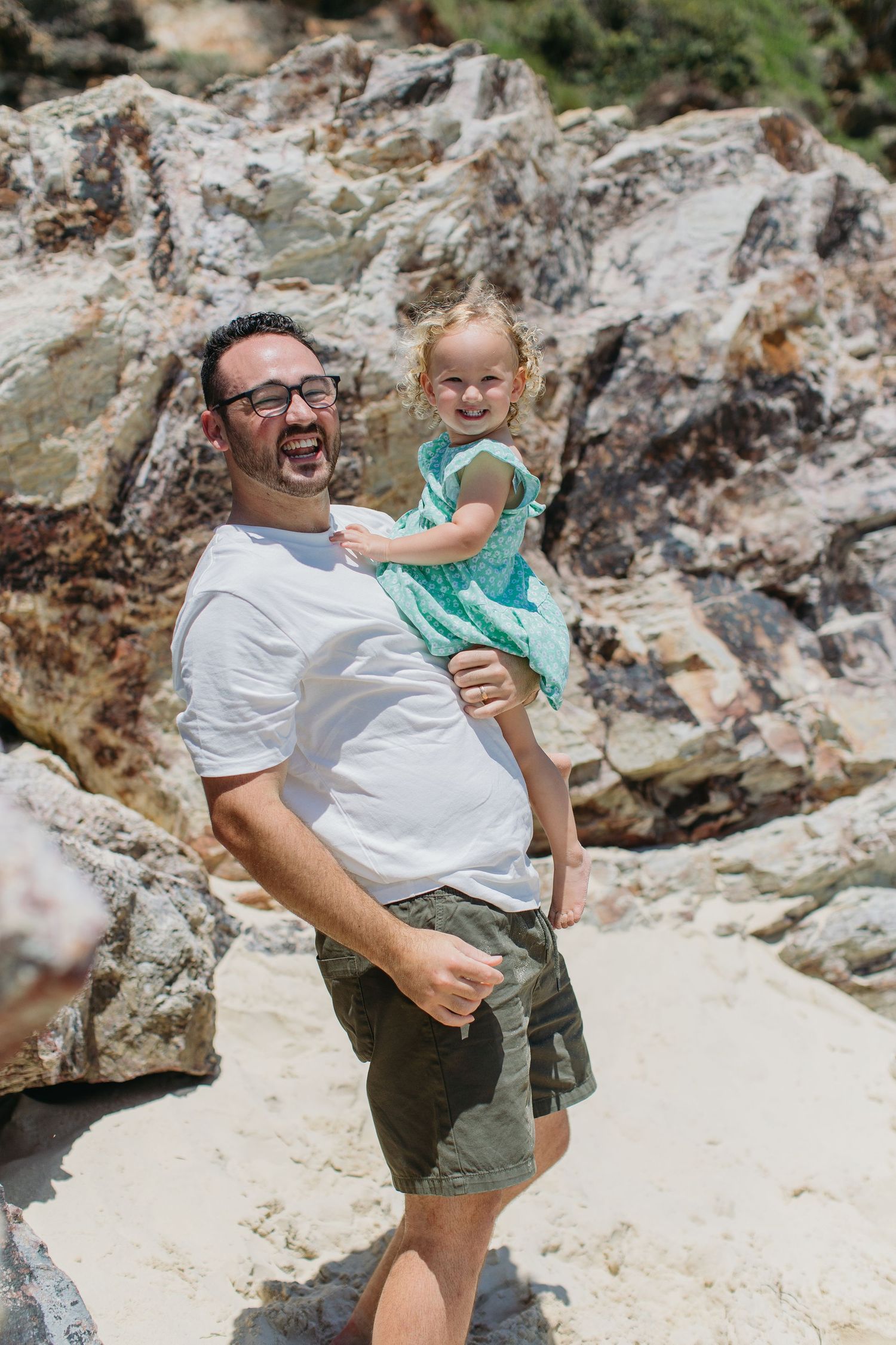 Laughing father in white shirt holds young girl in teal dress on sandy rocky beach.
