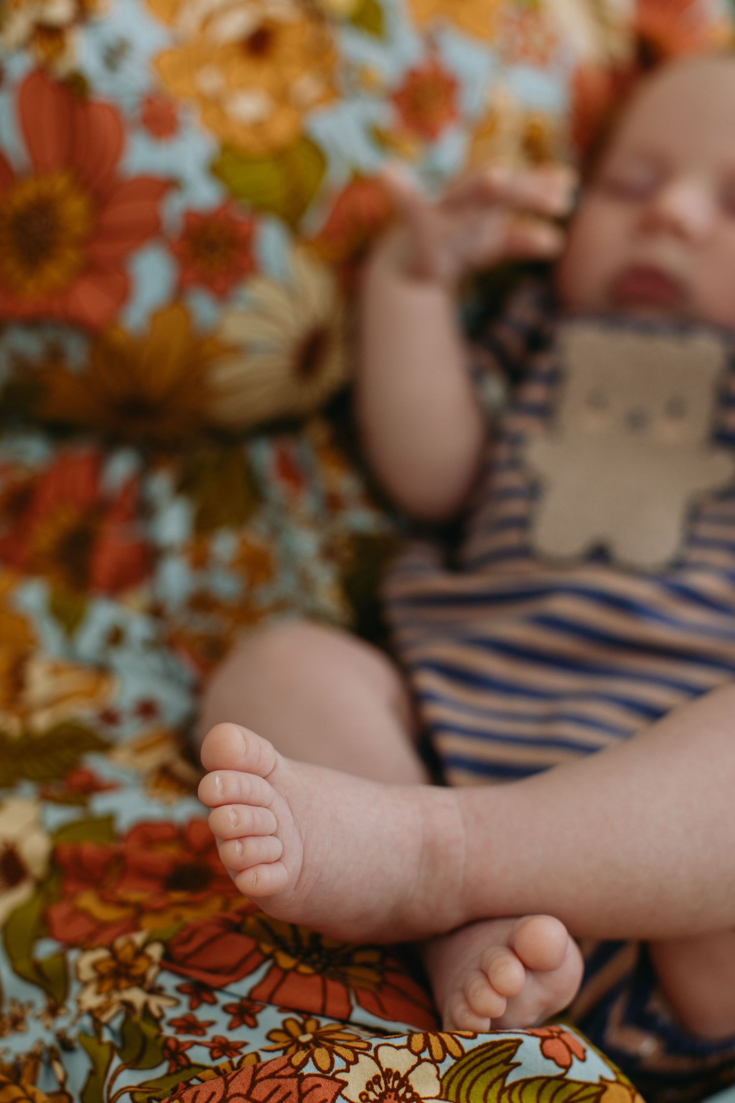 Newborn baby sleeping on a vintage floral blanket wearing a striped onesie with tiny bare feet in focus.
