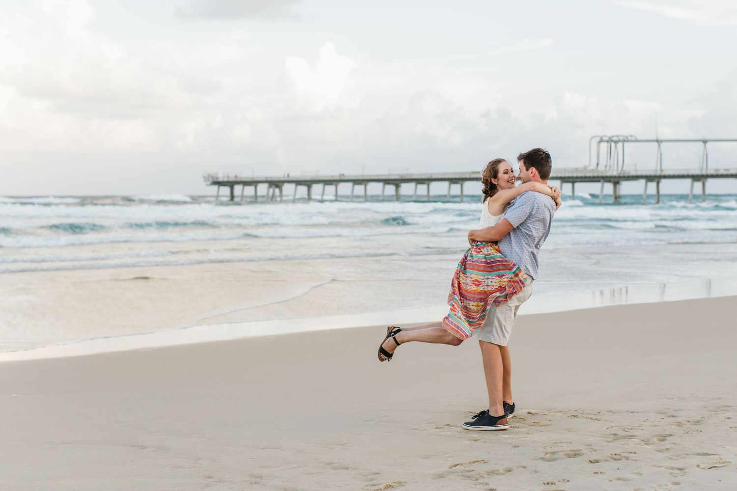 Couple embracing on a sandy beach near a pier, with ocean waves and cloudy sky in the background.