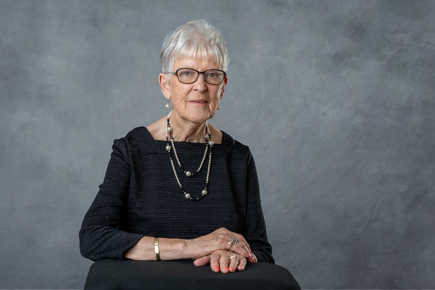 Elegant senior woman with short gray hair in black dress seated with hands folded.