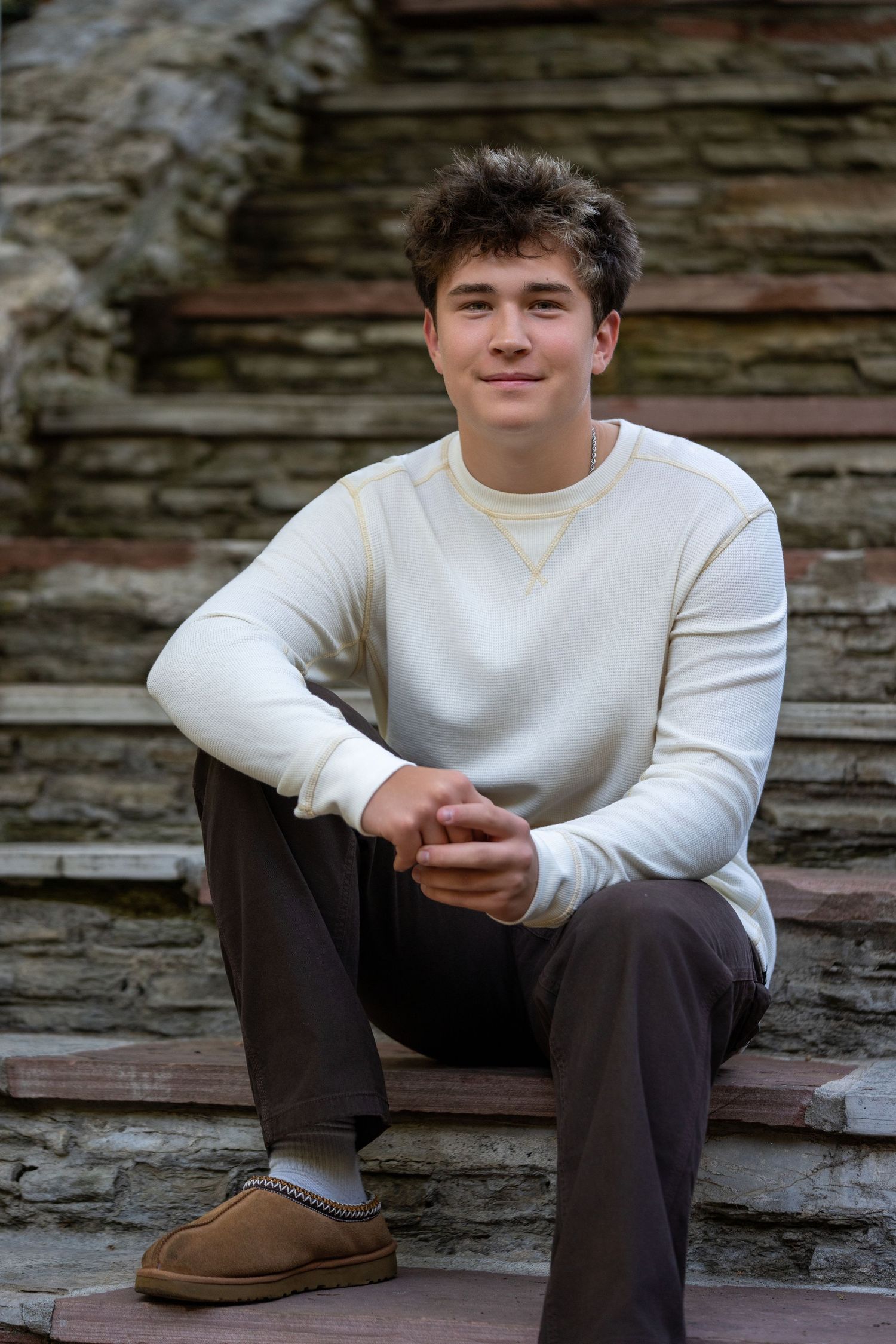Teen boy in white sweater and UGG slippers sitting on stone steps smiling at camera.