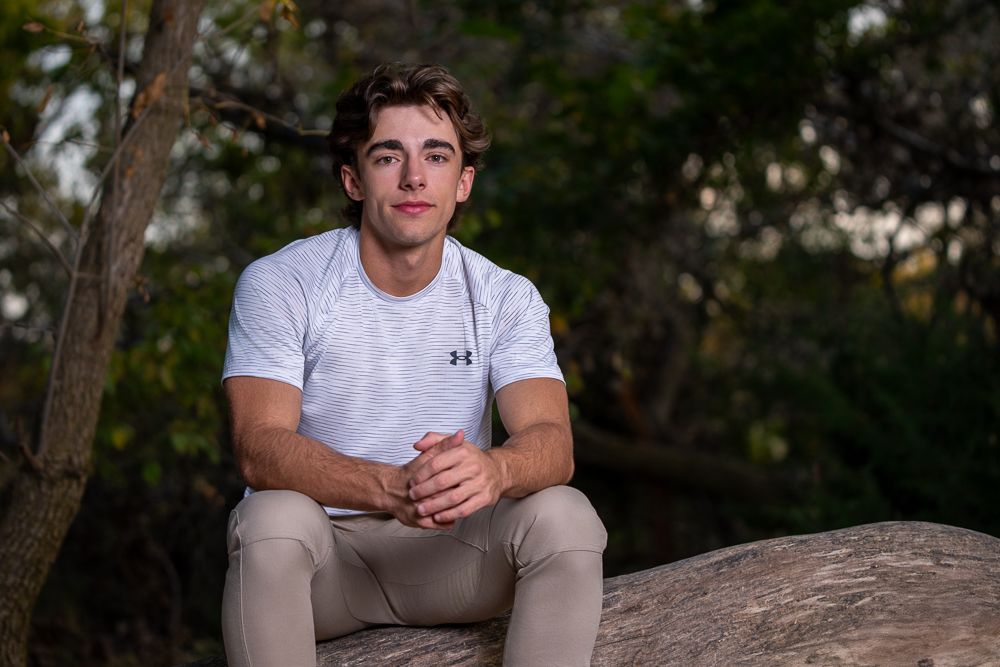Young man in white athletic shirt and khaki pants sitting on a rock outdoors with trees in background.