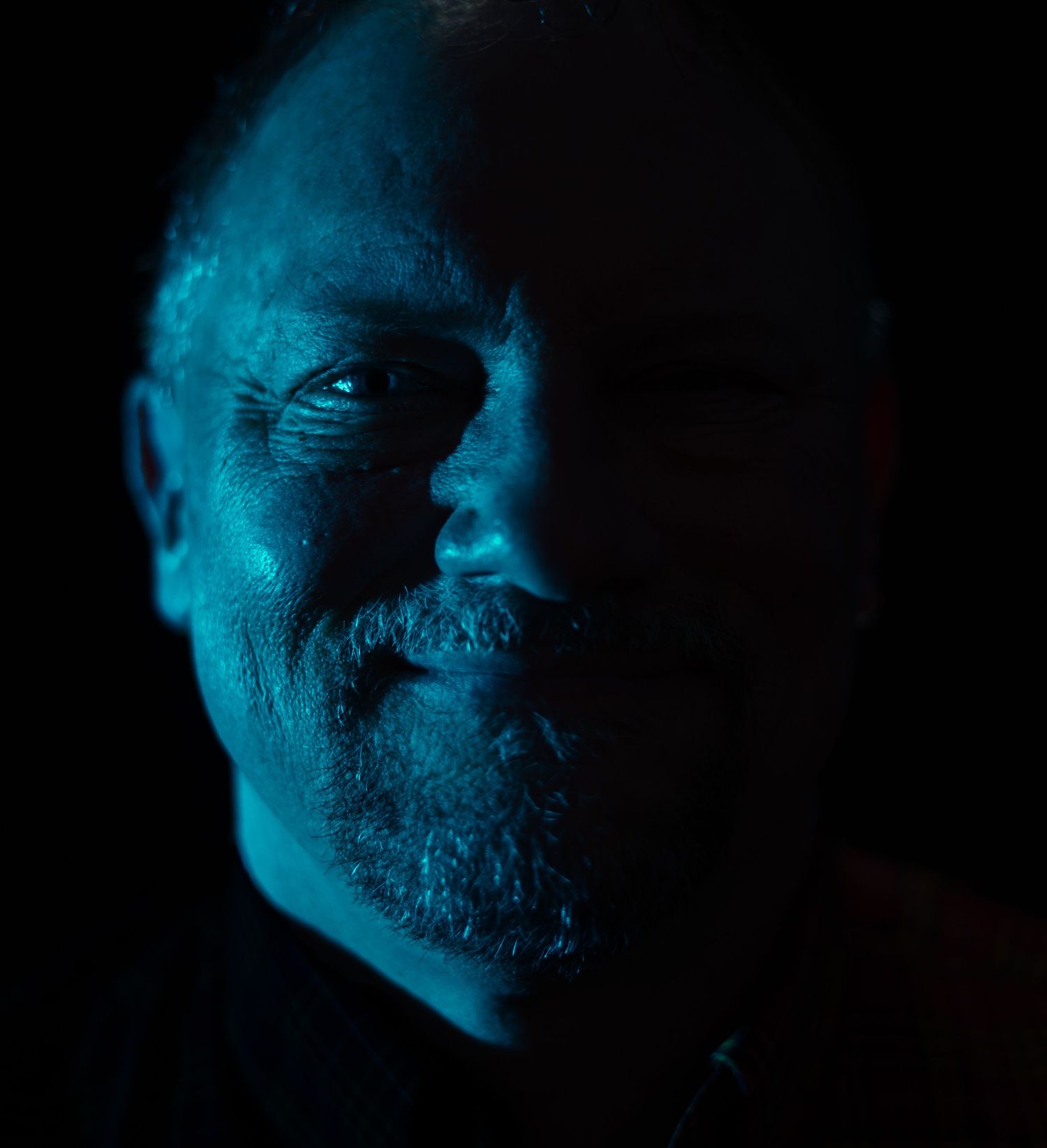 Dramatic blue-lit portrait of a man smiling in a dark, moody setting.