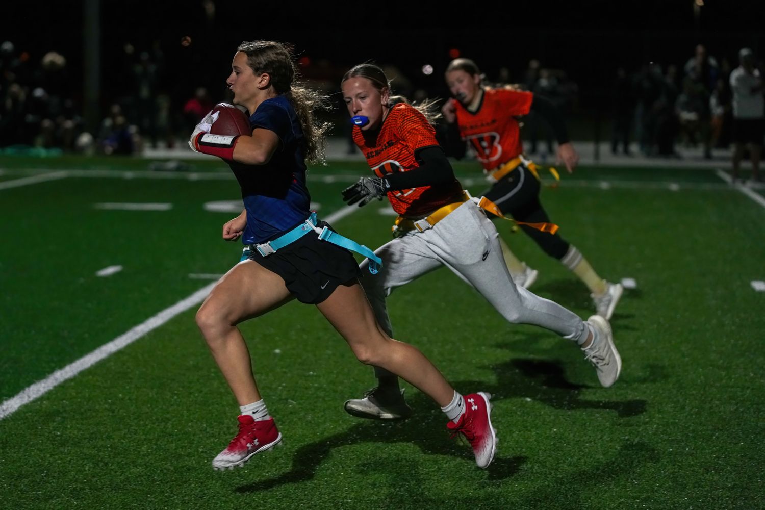Girls flag football players racing downfield at night under stadium lights.