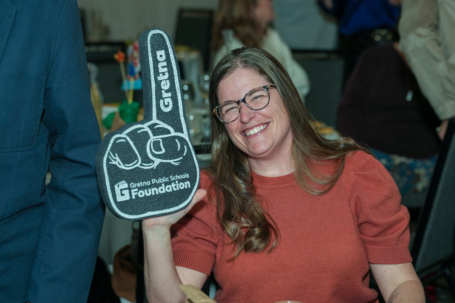Smiling woman holds a large Gretna Public Schools Foundation foam finger at a school fundraising event.