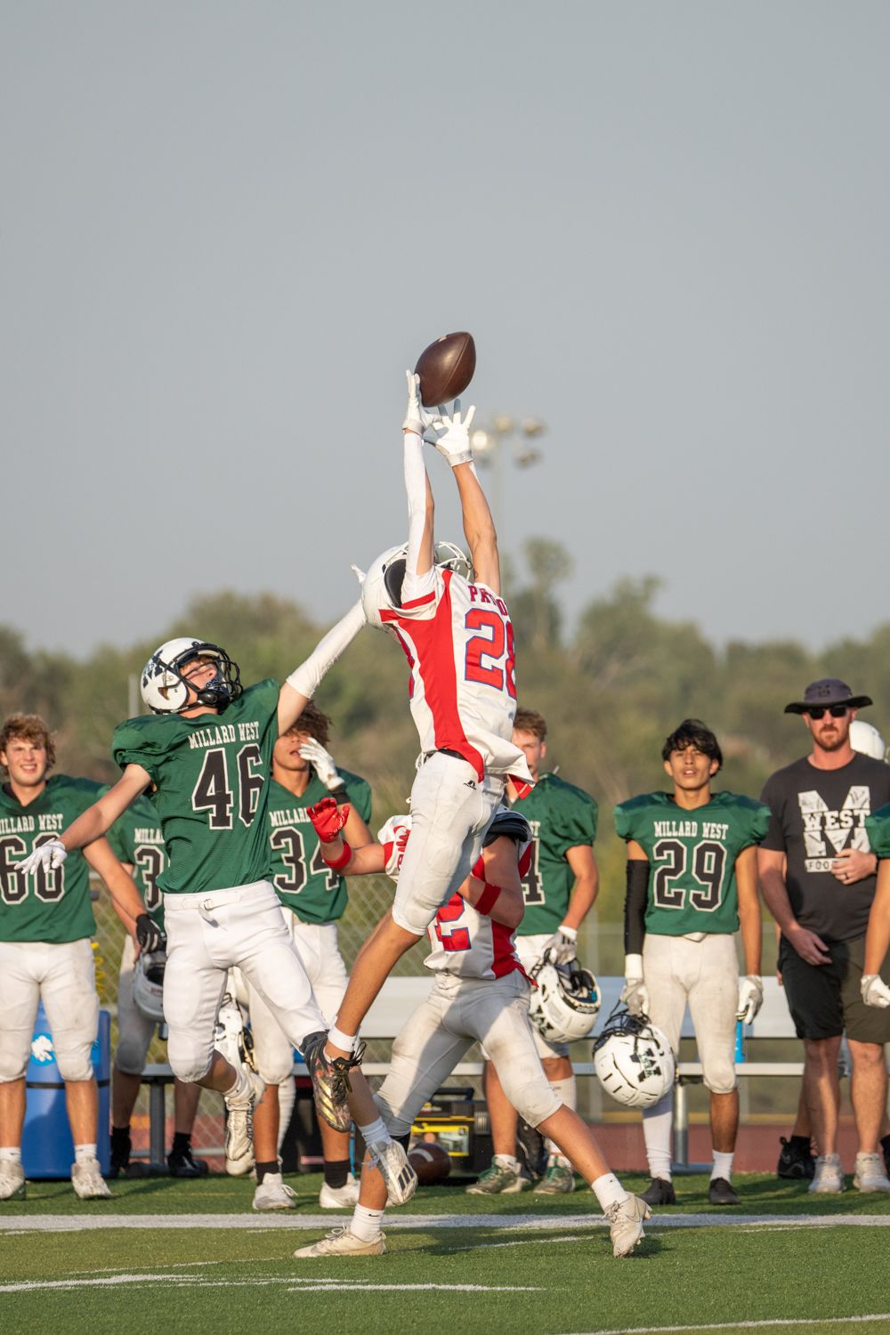 Football player #22 in red leaps high to catch a pass over defenders in green Millard West uniforms during a game.