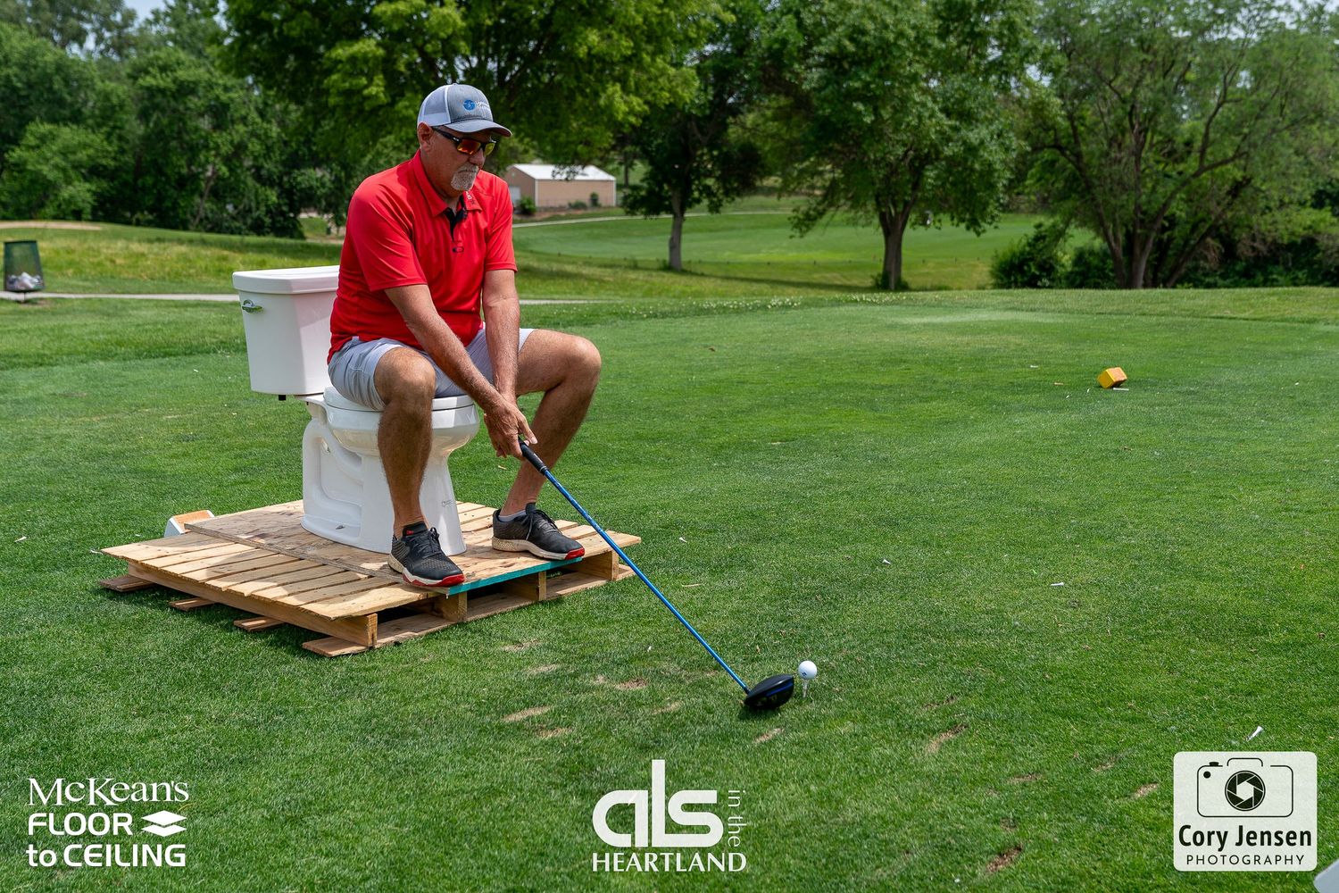 Golfer sits on a toilet on a wooden pallet while taking a golf shot on a green course at ALS charity event.