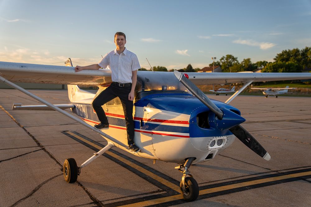 Young pilot in white shirt stands confidently on a blue and white small aircraft on a sunny airfield.