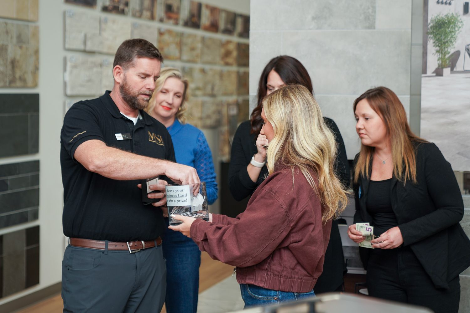 Man in black polo shirt drawing for winners with a group of women at an indoor event.