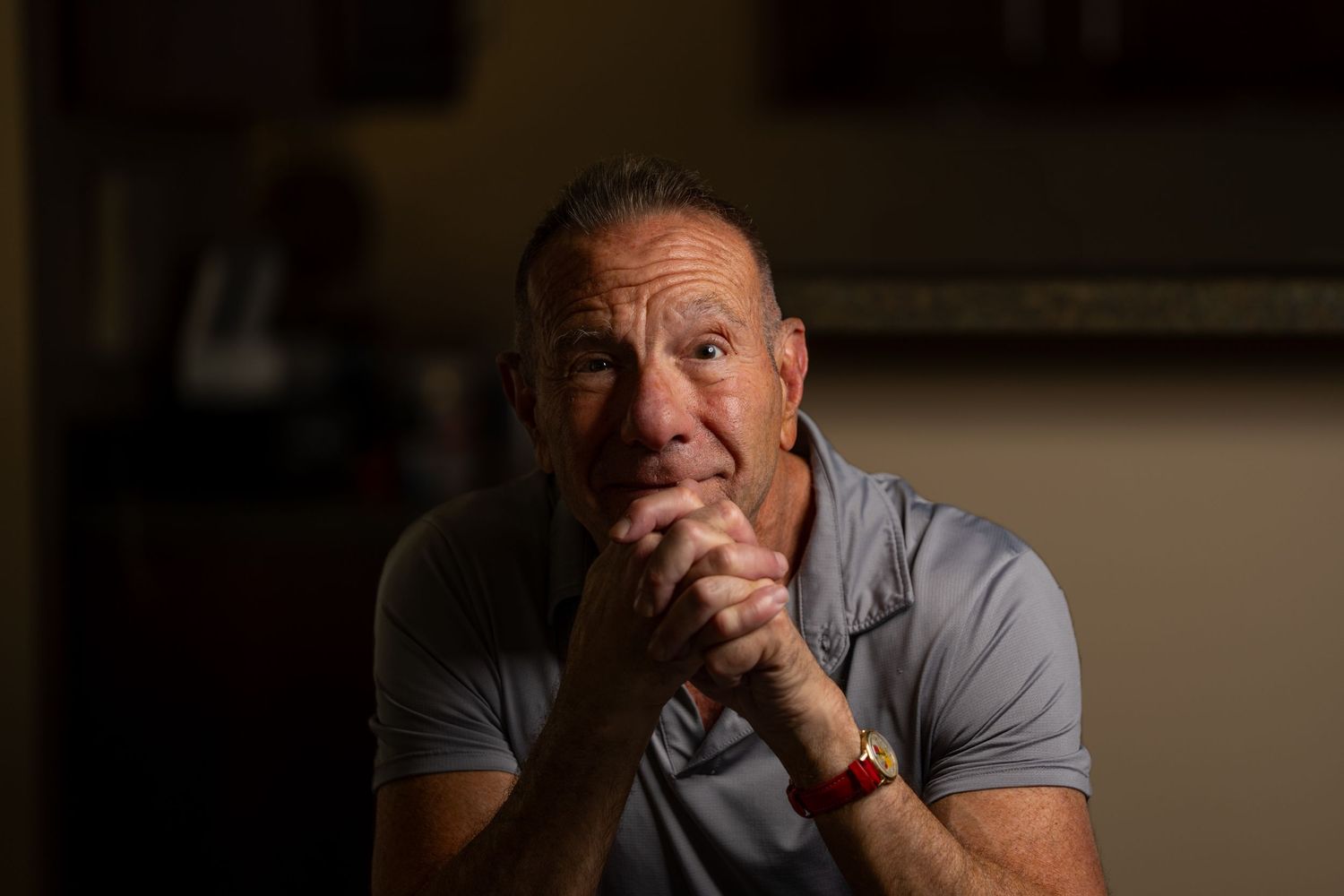 Middle-aged man with clasped hands sitting in a dimly lit room with a serious, contemplative expression.