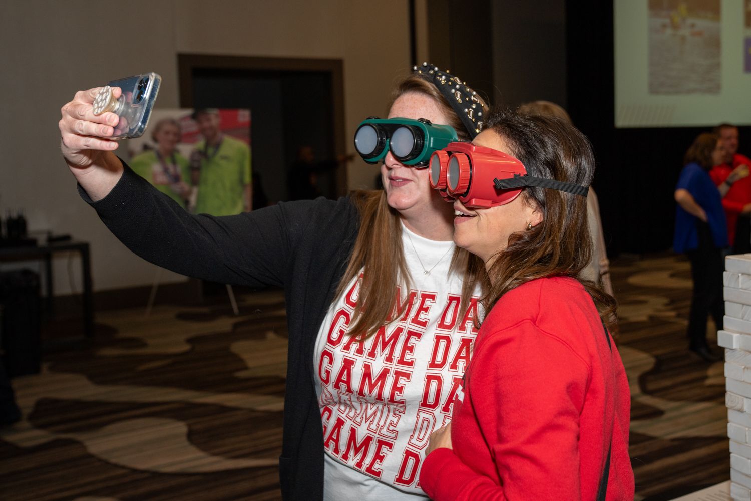 Two young women wearing novelty goggles taking a fun selfie together at an indoor event or conference.