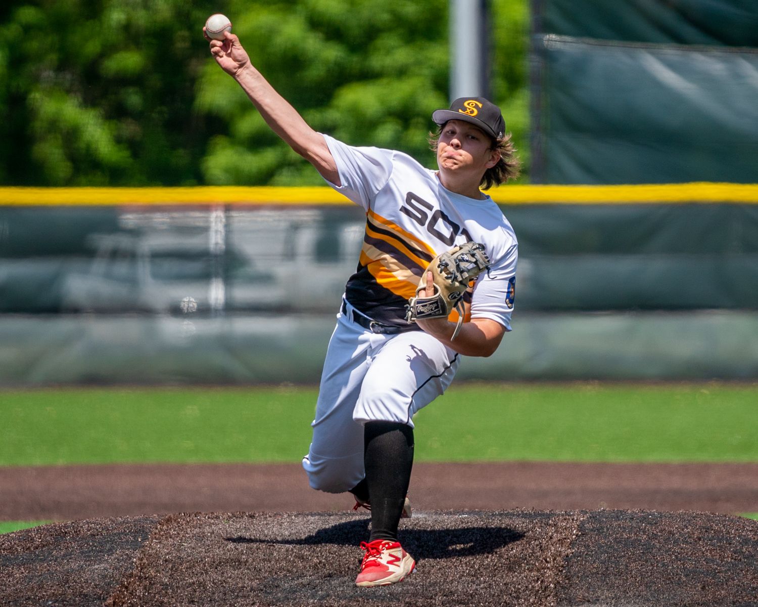 Teen baseball pitcher in gold and white uniform winds up to throw during game.