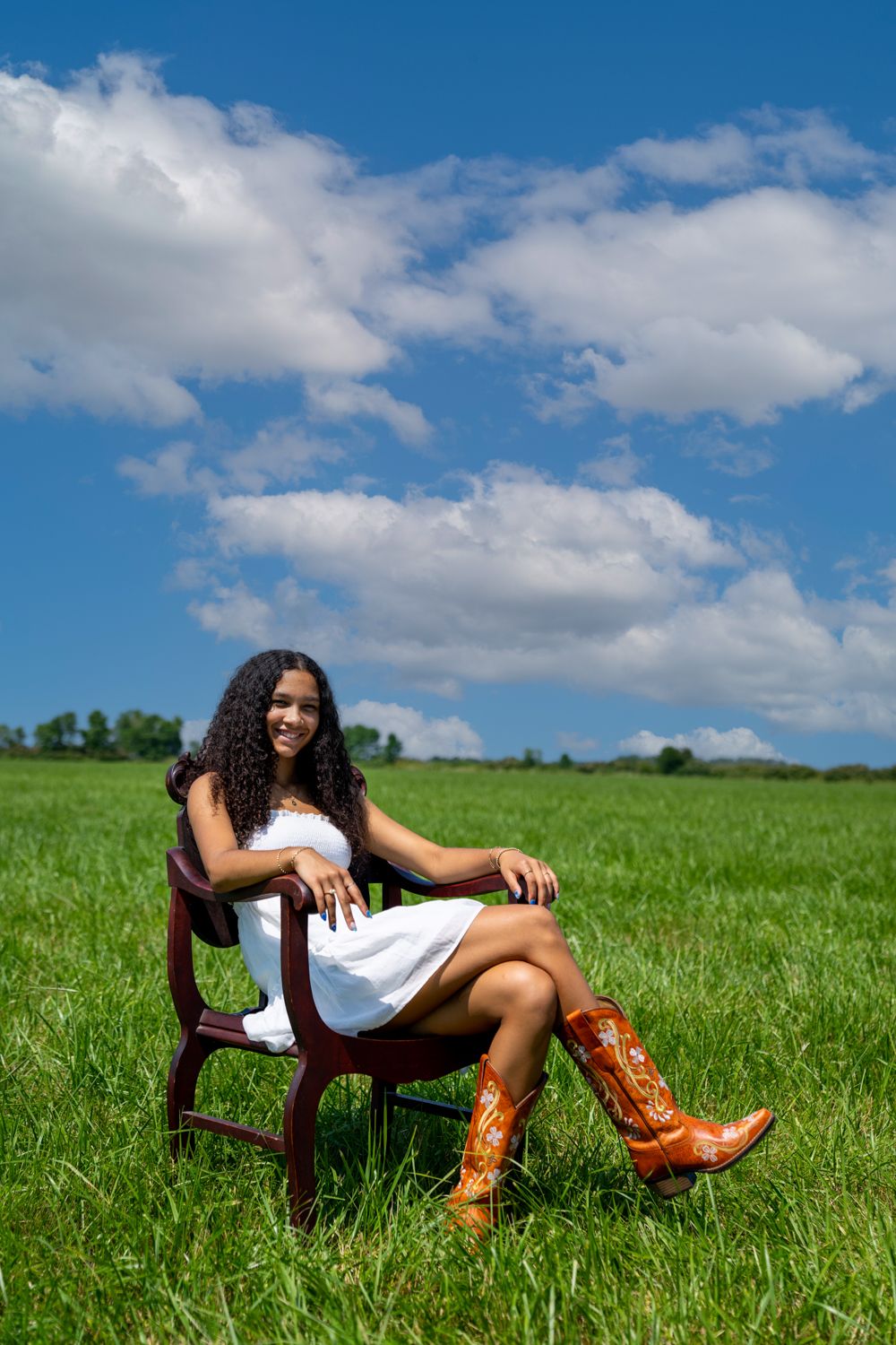 Young woman in white dress and cowboy boots sitting in wooden chair in a green meadow under blue sky.