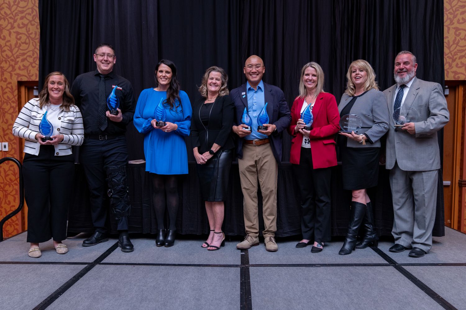 Group of eight professionals smile and pose with blue awards on a stage at a formal recognition ceremony.