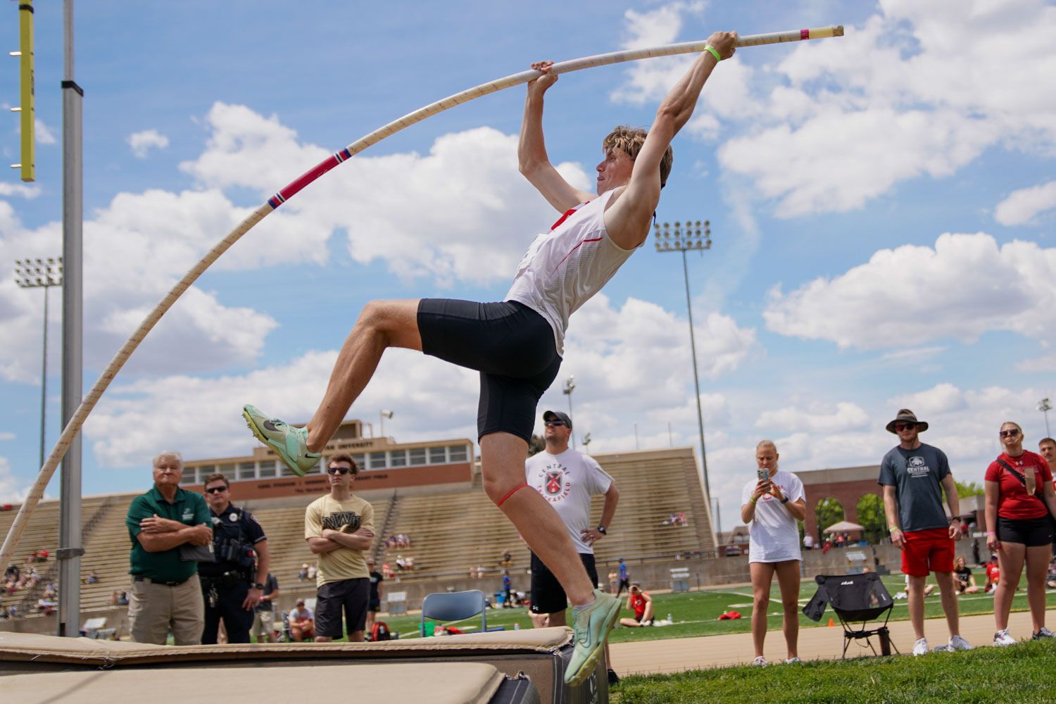 Male athlete performing a pole vault at an outdoor track and field meet with spectators watching in the background.