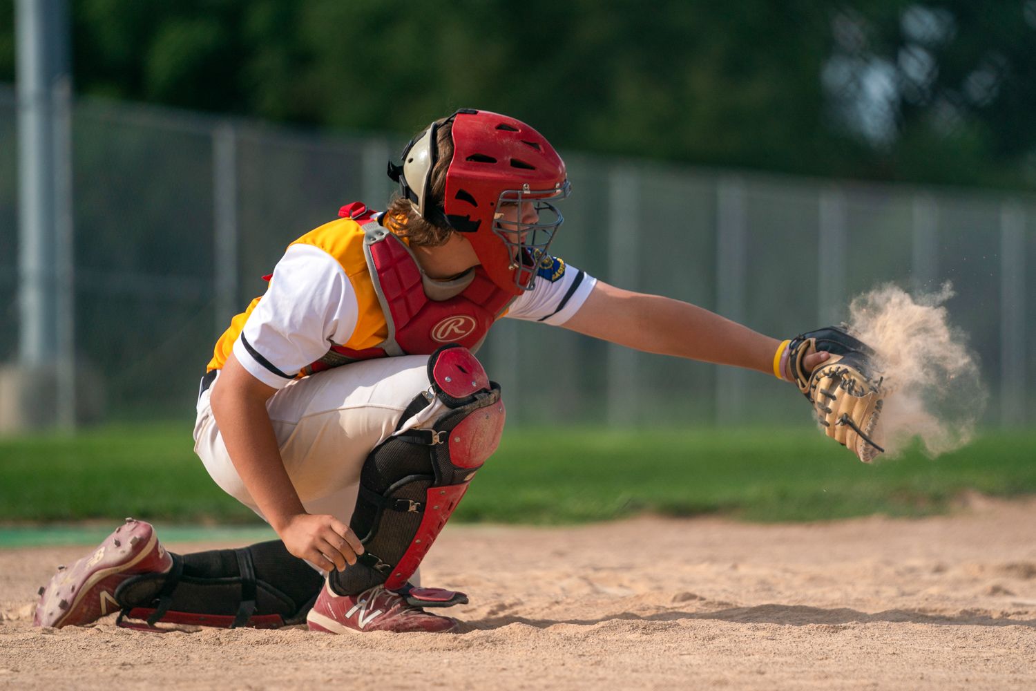 Baseball catcher in red helmet and gear crouches at home plate catching a pitch in a cloud of dust.